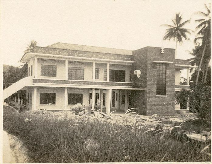 Black and white photo of a two-story building with a brick section, surrounded by tall grass and palm trees.
